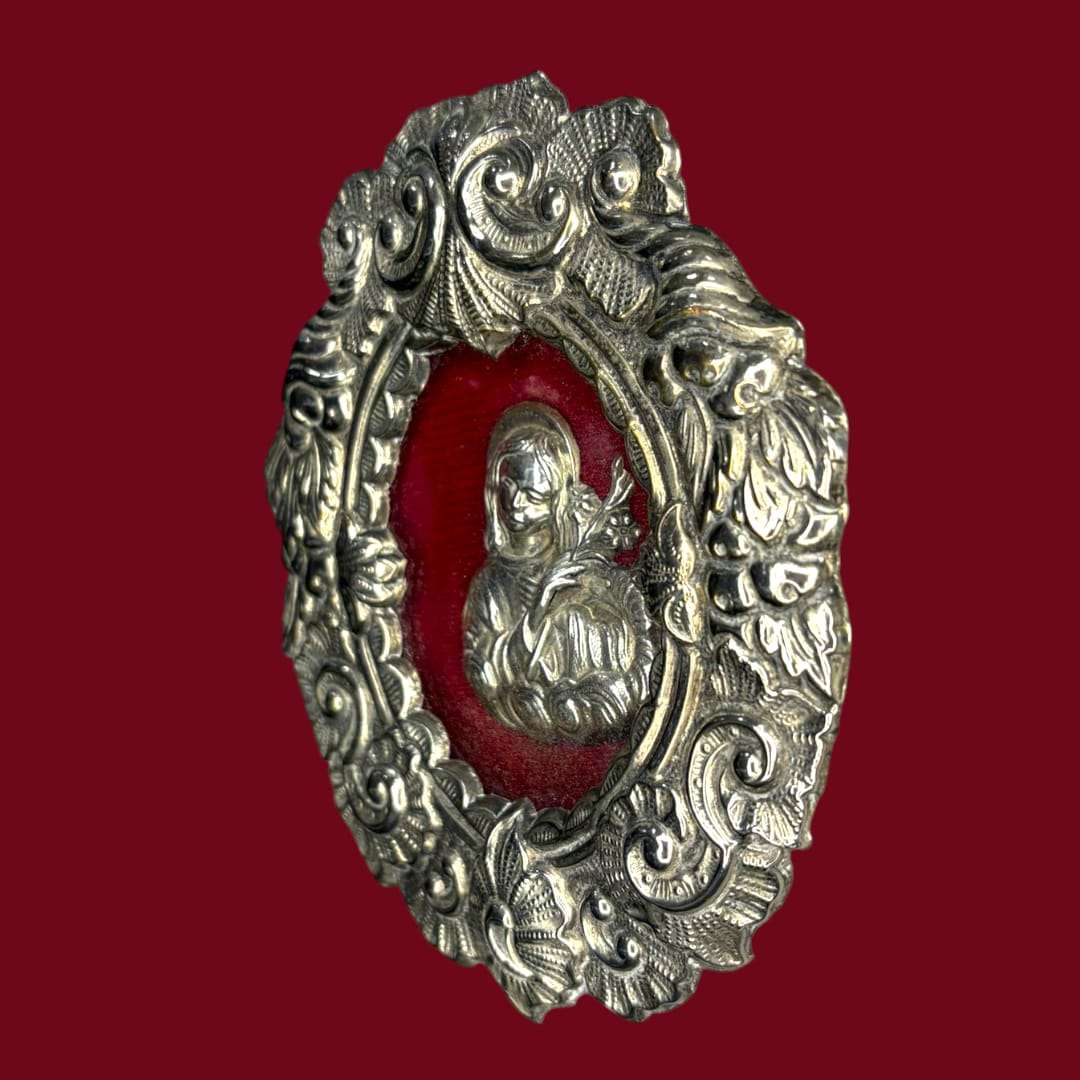 Se trata de una pieza de arte religioso tradicional elaborada con un marco de metal ricamente ornamentado y una imagen en relieve sobre un fondo rojo intenso que realza su presencia espiritual.