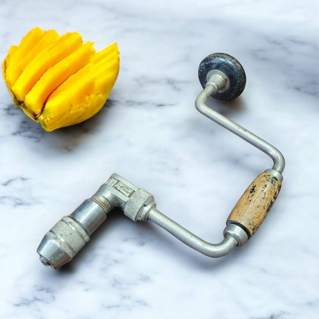 Taladro manual vintage de manivela con mango de madera sobre fondo de mármol junto a una fruta amarilla. Ideal para decoración rústica.