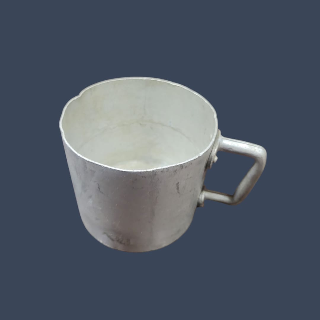 Taza de aluminio con mango junto a una cafetera vintage, creando una escena retro.