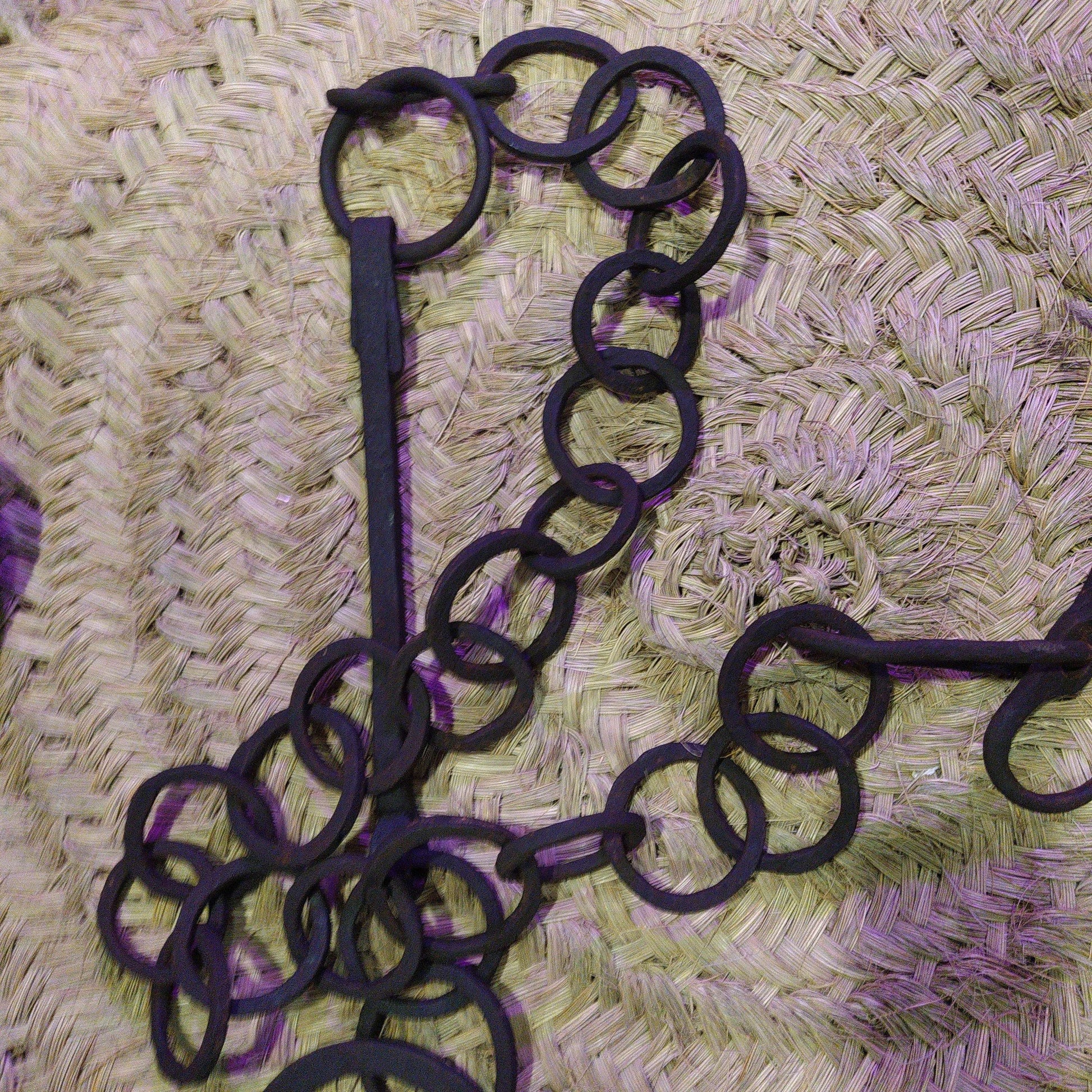 Cadena forjada de hierro con eslabones circulares sobre fondo de mimbre, ideal para colgar calderos en chimeneas rústicas.