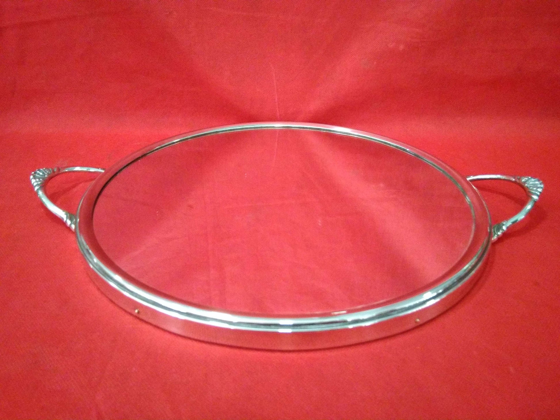 Bandeja redondeada de metal plateado espejada con asas decorativas sobre fondo rojo. Diseño elegante ideal para decoración o servir.