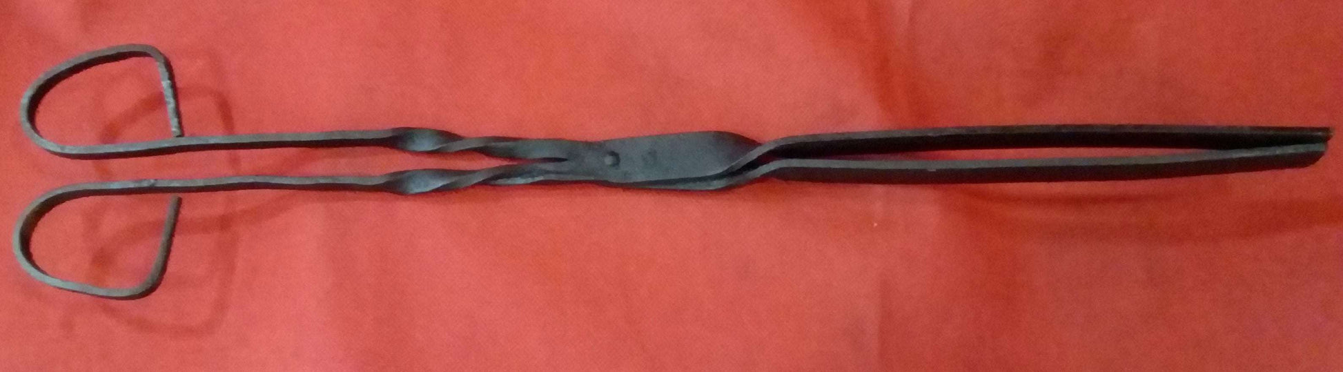 Pinza para brasero en hierro forjado con diseño rústico sobre fondo rojo. Ideal para decoración vintage en hogar y jardín.
