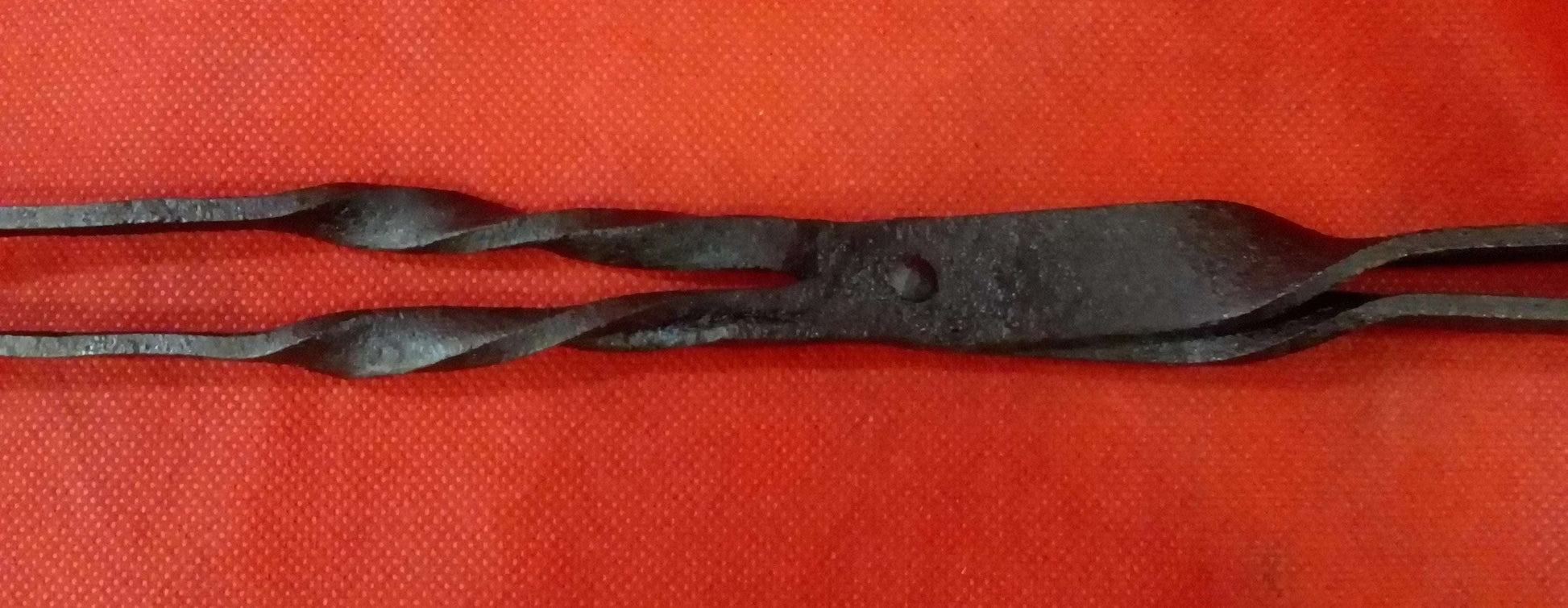 Pinza para brasero en hierro forjado con diseño rústico sobre fondo rojo. Ideal para decoración vintage en hogar o jardín.