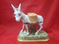 Figura de burro de porcelana con alforjas sobre base decorativa, ideal para decoración vintage. Disponible en Cositas Antiguas.