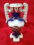 Jarrón de cerámica pintado a mano con asas ornamentales y diseño exclusivo de casa rural sobre fondo azul. Ideal para decoración elegante y antigua.