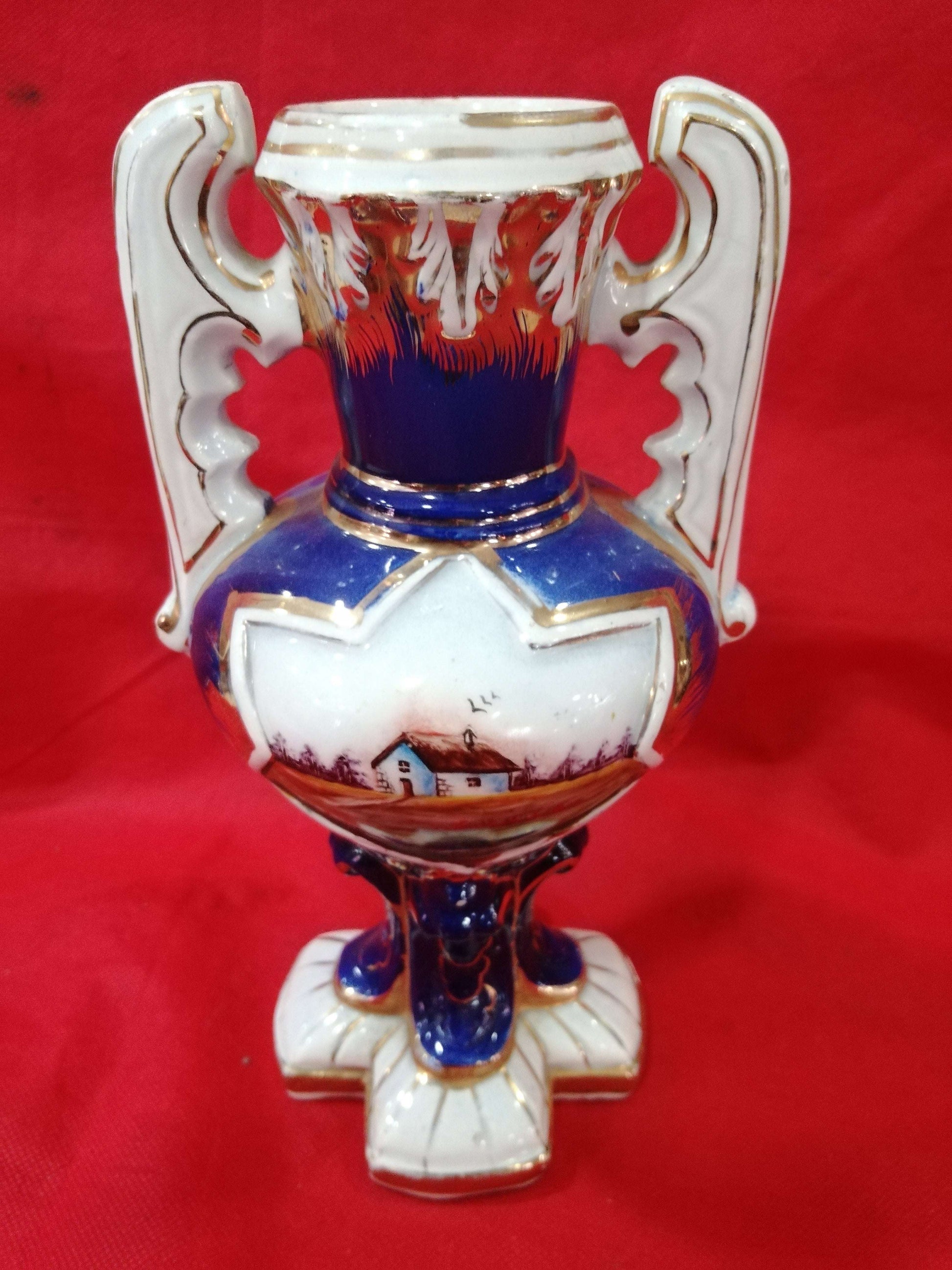 Jarrón de cerámica pintado a mano con asas ornamentales y diseño exclusivo de casa rural sobre fondo azul. Ideal para decoración elegante y antigua.