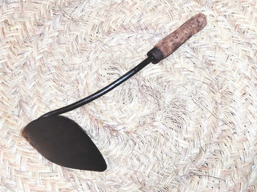 Amocafre con mango de madera curvo sobre fondo de palma tejida. Herramienta de jardín única ideal para decoración rústica o uso práctico.