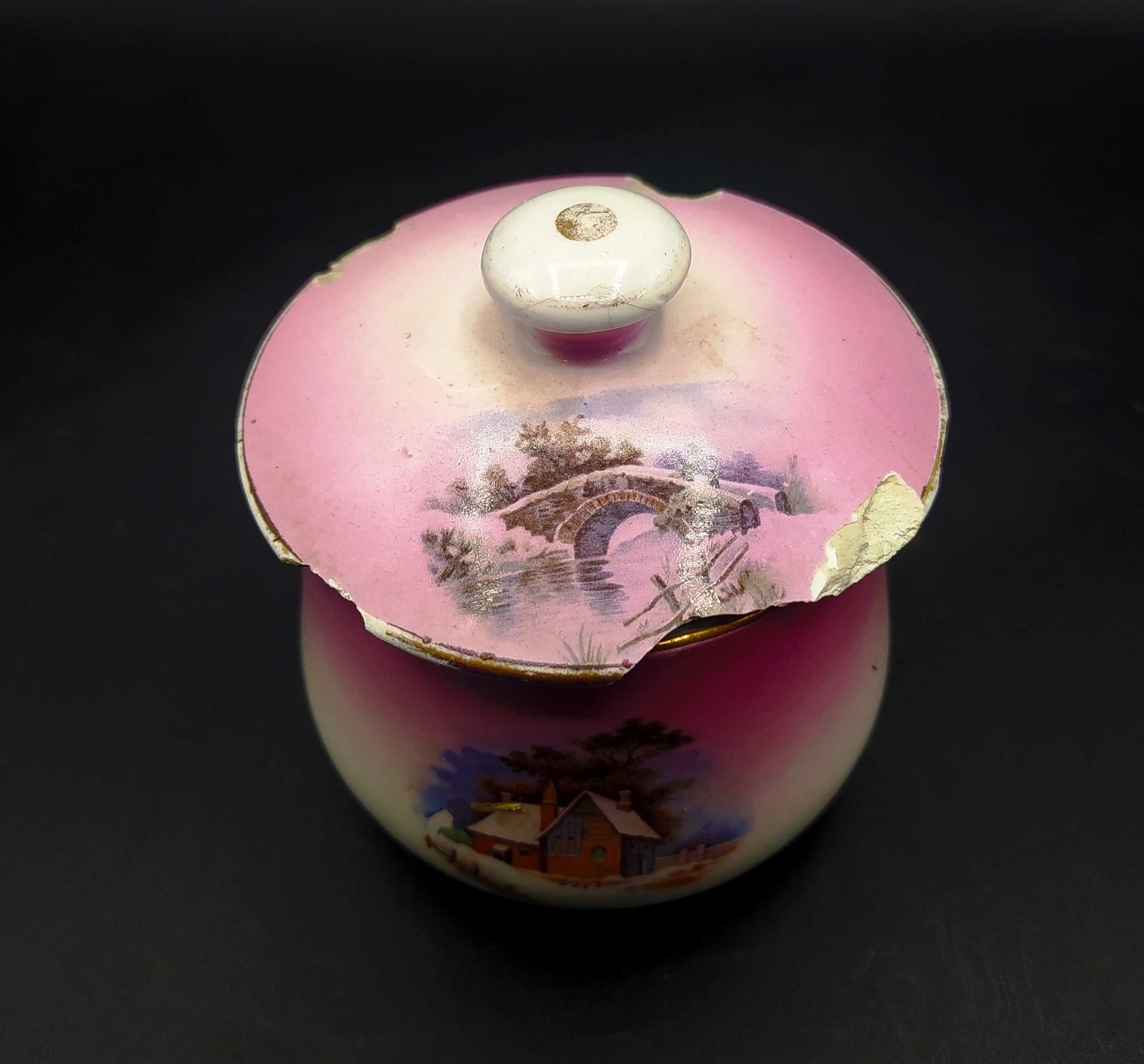 Tarro de tocador en porcelana rosa de Cartuja de Sevilla con tapa decorada con paisaje y bordes desgastados. Ideal para decoración vintage.