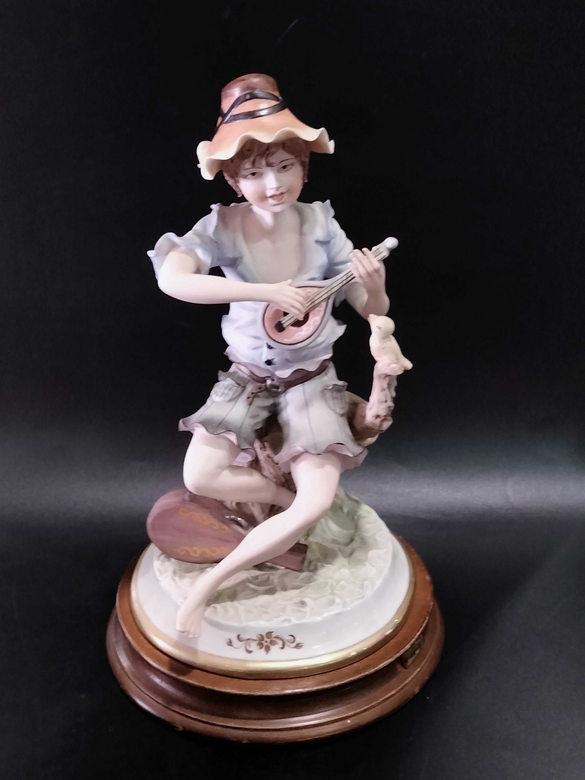 Figura de porcelana Carlos de niño con sombrero tocando instrumento de cuerda, sentado sobre base de madera, pieza decorativa de colección.