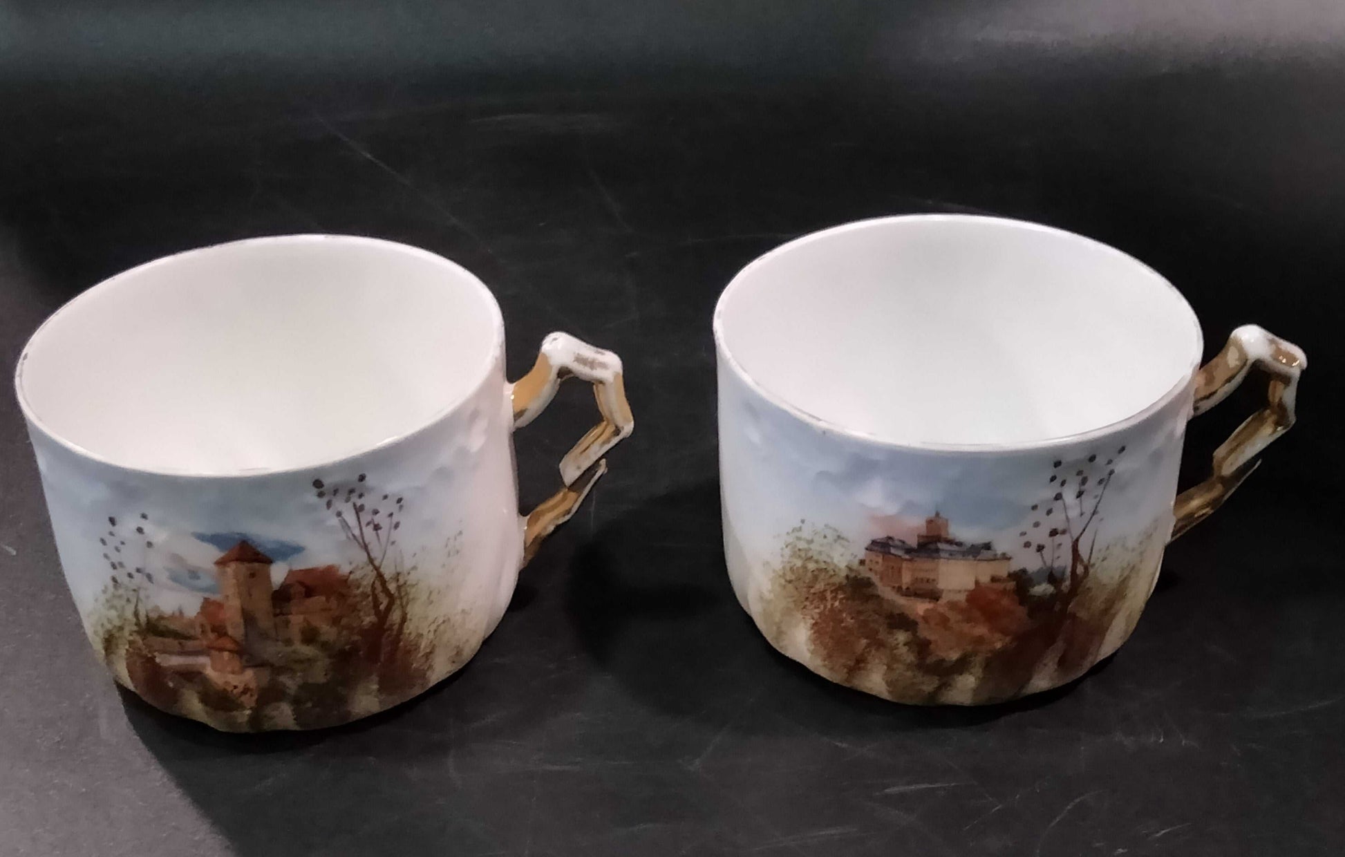 Par de tazas de porcelana Limoges del Juego de Café Tú y Yo, con asas doradas y paisajes pintados a mano, ideales para decoración o coleccionismo.