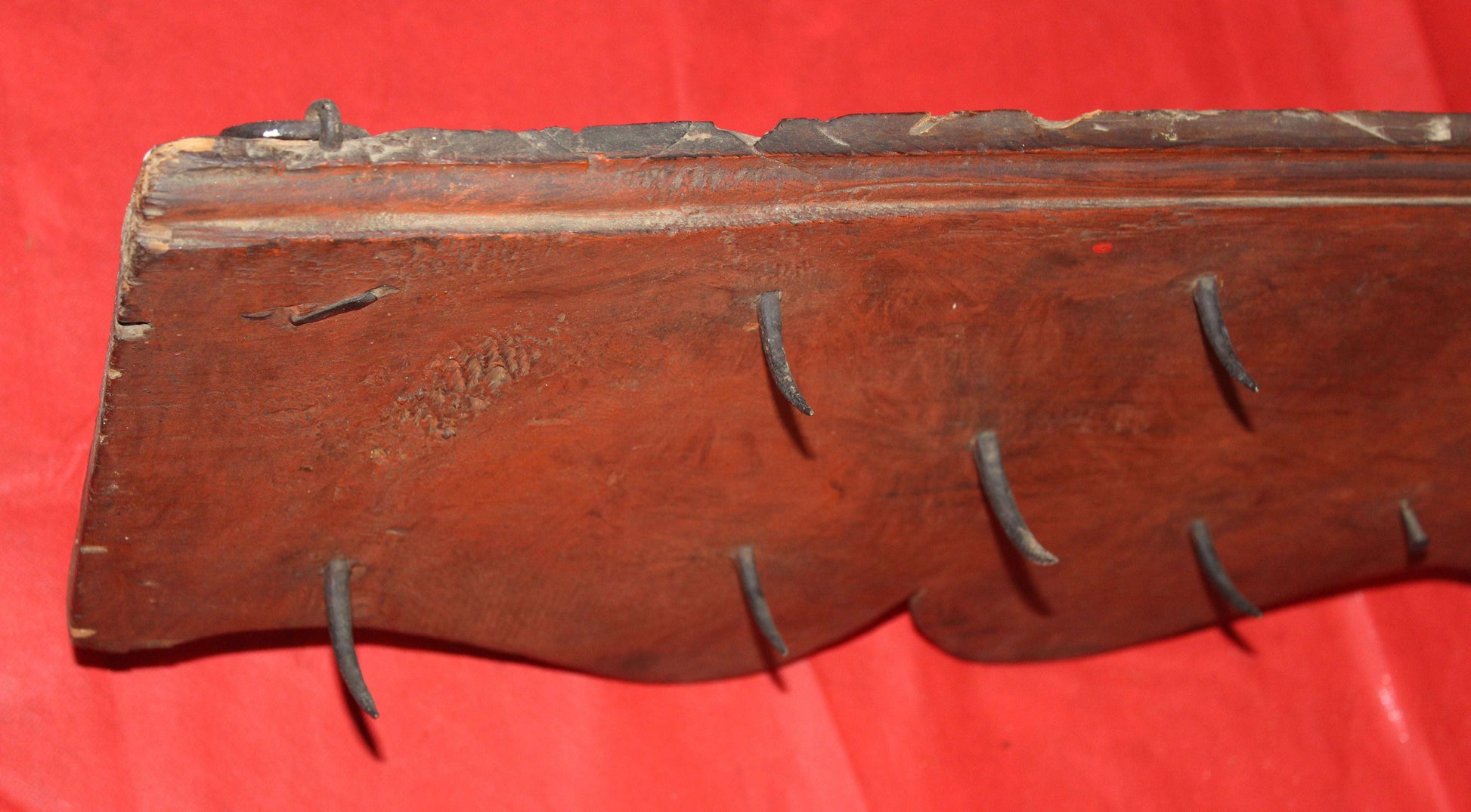 Percha antigua de matanzas con clavos de fragua, estilo rústico, en madera oscura y ganchos forjados a mano sobre fondo rojo. Ideal para decoración vintage.