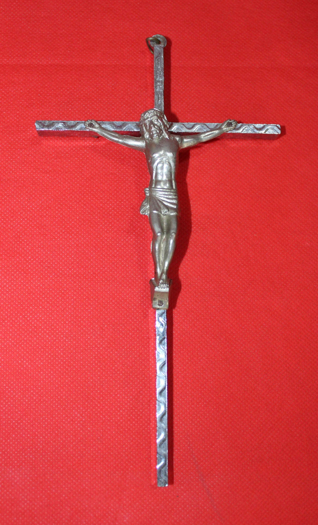 Crucificado de metal plateado sobre fondo rojo, diseño artesanal con detalles en relieve. Decoración religiosa de calidad y tradición cristiana.