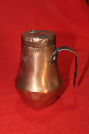 Jarra antigua de cobre con tapadera sobre fondo rojo, ideal para decoración vintage en hogar y jardín. Diseño rústico con asa metálica artesanal.